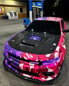 a pink camo car is parked at a gas station with its hood up and it's lights on