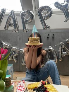 a woman sitting at a table with a cake in front of her and balloons behind her