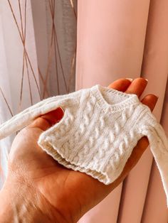 a hand holding a white knitted sweater in front of a window with pink curtains