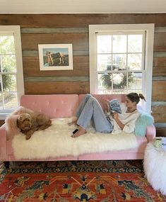 a woman sitting on a pink couch reading a book with a dog laying next to her