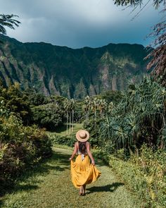 a woman in a yellow dress and straw hat walks down a path with mountains in the background