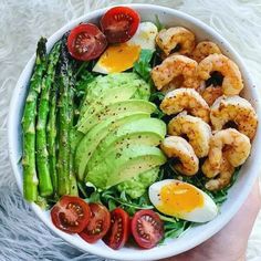 a person holding a white bowl filled with shrimp, avocado, tomatoes and asparagus