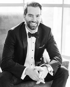 a man in a tuxedo is sitting on a couch and smiling at the camera