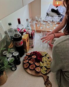 a woman standing in front of a table filled with bottles and glasses next to an assortment of fruit