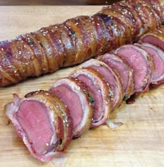 sliced meat on a wooden cutting board next to a large piece of meat wrapped in bacon