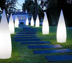 several tall white lights in the grass near a path leading to a building and trees