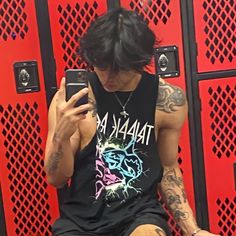 a man sitting in front of red lockers looking at his cell phone while wearing a tank top