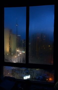 a window with rain falling on it and the city lights seen from outside at night