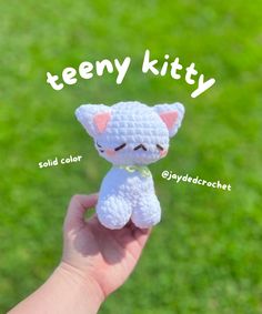 a hand holding a small white crochet kitty toy in front of green grass