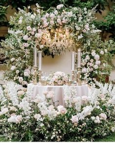 a table with white flowers and candles on it in front of a wall full of greenery