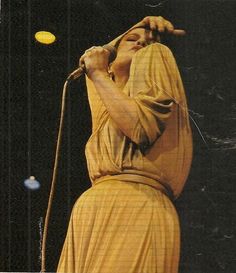 a woman in a yellow dress holding a microphone up to her ear and singing into the air