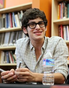 a young man sitting at a table in front of a book shelf holding a cell phone