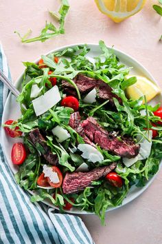 a salad with steak, tomatoes and cheese on it in a white bowl next to a lemon wedge