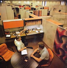 two people sitting at a table in an office cubicle with lots of desks