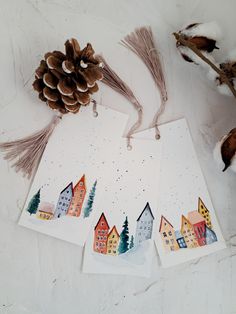 three cards with watercolor houses and pine cones on them next to some dried branches