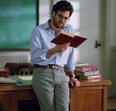 a man reading a book while standing in front of a desk with books on it