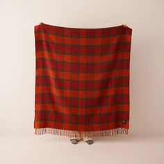 a red and black plaid blanket with fringes on it's end, in front of a white wall