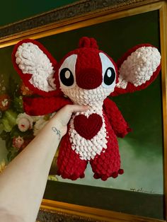 a hand holding a red and white crocheted stuffed animal in front of a painting