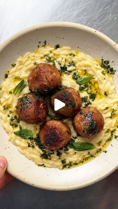 a white bowl filled with pasta and meatballs on top of a metal table next to a person's hand