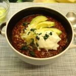 a bowl of chili with sour cream and broccoli next to a glass of wine