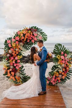 a bride and groom standing in front of an orange flower arch on the beach at sunset