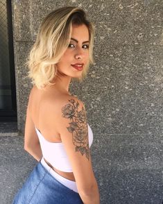 a woman with a tattoo on her arm standing in front of a wall and looking at the camera