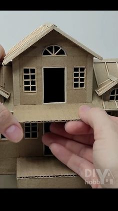 two hands are holding a model of a house with windows and shutters on it