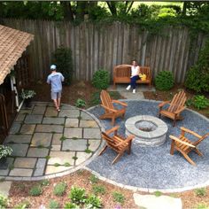 a backyard with a fire pit surrounded by wooden chairs and benches, as well as two people walking in the back yard