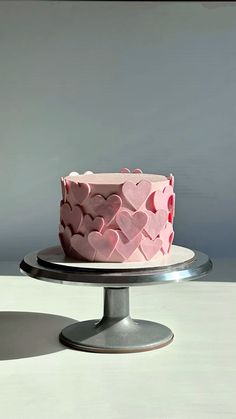 a pink cake with hearts on it sitting on top of a metal stand in front of a gray wall