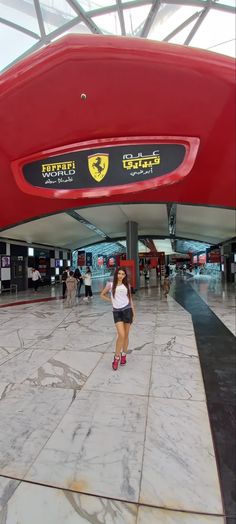 a woman is standing in front of a large red car at the ferrari museum, with people walking around