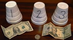 three stacks of money sitting on top of a wooden table next to two cups and one dollar