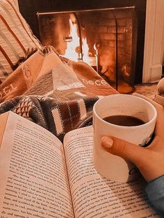 a person holding a cup of coffee while reading a book in front of a fireplace