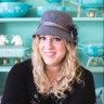 a woman wearing a hat sitting in front of a shelf filled with blue and green dishes