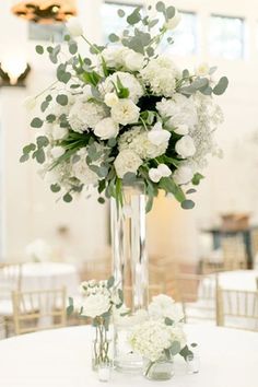 a tall vase filled with white flowers and greenery on top of a round table