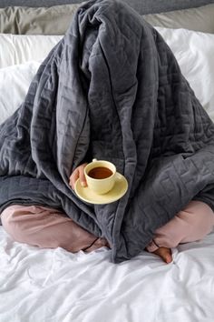 a person sitting on a bed with a blanket over their head and holding a cup of coffee