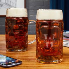 two beer mugs sitting next to each other on top of a wooden table near a cell phone