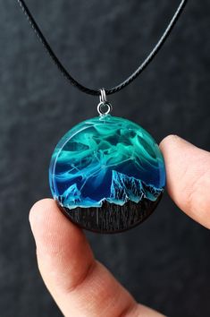 a hand holding a glass pendant with mountains and water in the background on a black cord