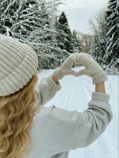 a woman is making a heart shape with her hands while standing in front of snow covered trees