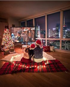 two people sitting on the floor in front of a christmas tree watching tv at night