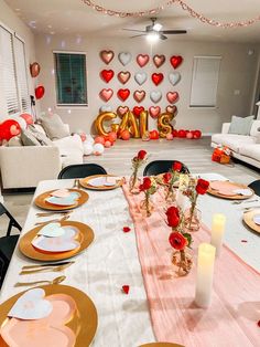 a table set for valentine's day with red roses and heart balloons on the wall