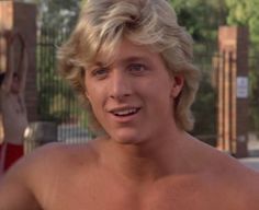 a man with blonde hair and no shirt smiling at the camera while standing in front of a fence