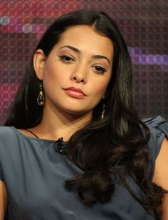 a woman with long dark hair and earrings sitting in front of a purple wall wearing a blue dress