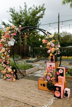 an outdoor area with flowers and speakers on the ground next to a circular arch that has lights strung from it