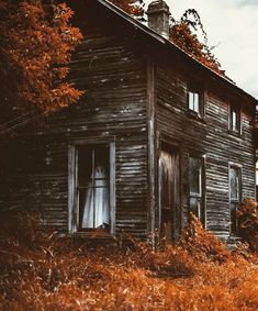 an old wooden house sitting in the middle of a field with tall grass and trees around it