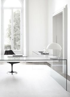 a glass table with an oval base in front of a white wall and large window