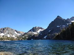 the mountains are covered in snow and trees by the water with clear blue skies above them