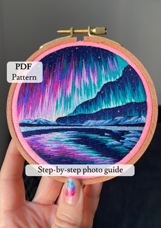 a person holding up a small embroidery project with an aurora bore in the sky on it