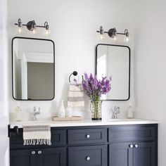 a white and black bathroom with two mirrors on the wall, purple flowers in vase