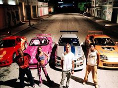 three men and two women standing in front of four different colored cars on the street