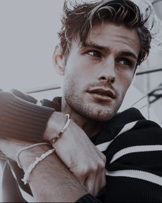 a young man wearing a sweater and bracelet with his hand on his arm, looking at the camera